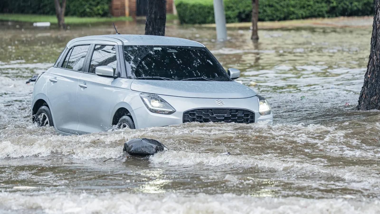 A silver hatchback driving through deep floodwater on a street
