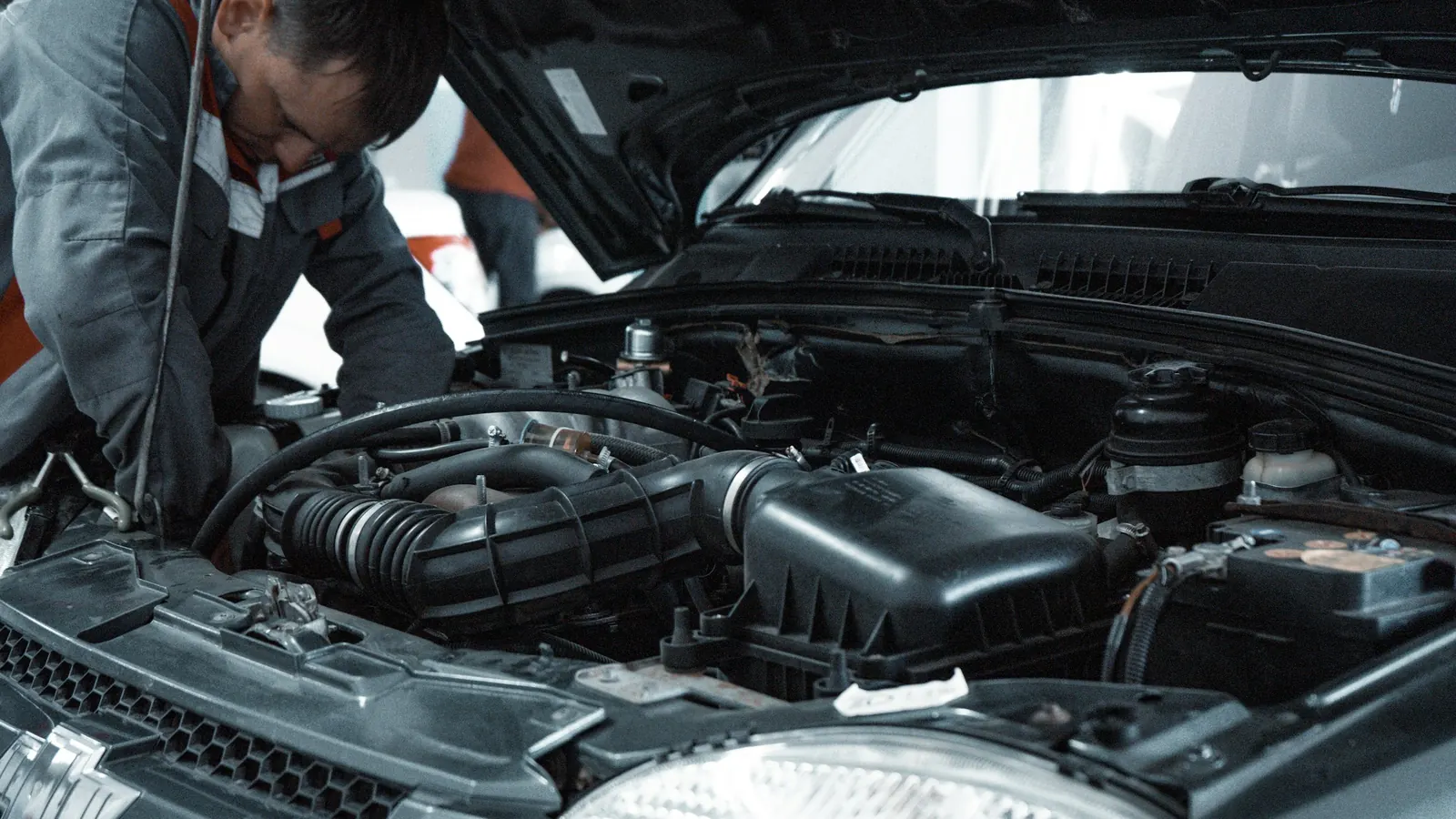 A mechanic in a grey uniform leans over an open engine bay to inspect the hoses and engine