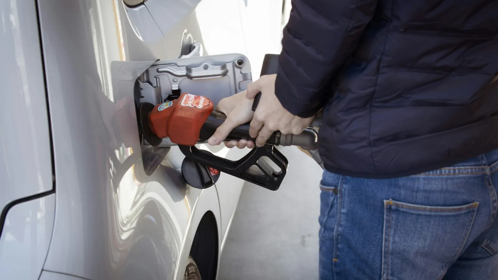 A driver filling a silver car with gas, the fuel door open and the black gas cap hanging on its tether